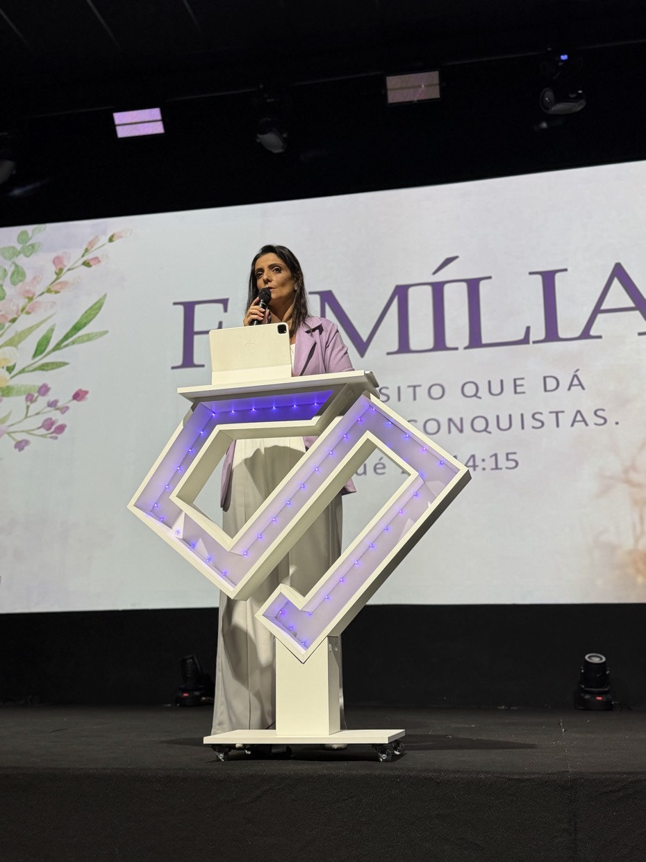 Adriana Ricci no púlpito diante de um telão com a palavra Família e a frase o propósito que dá sentido às conquistas.