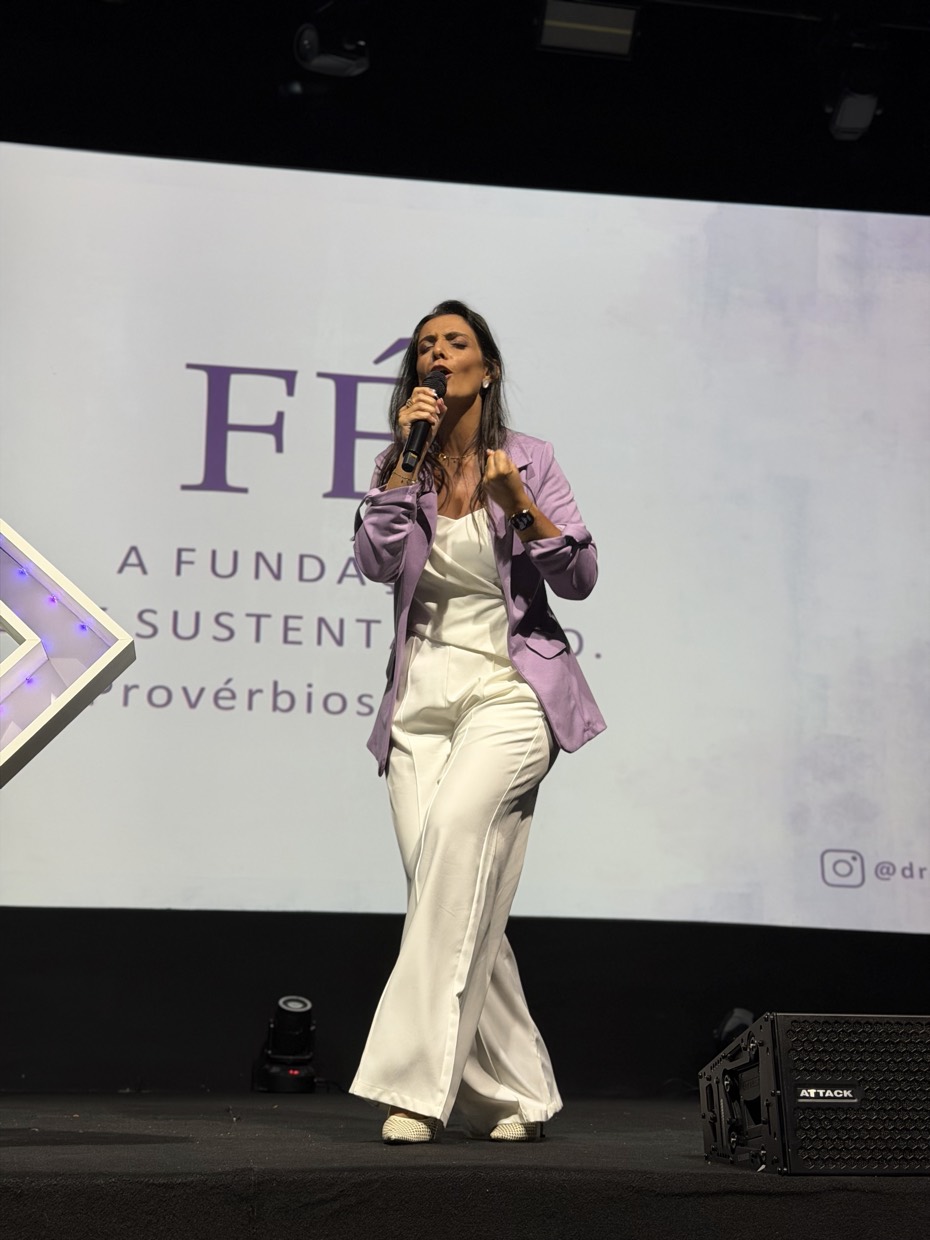 Adriana Ricci no palco, ao lado de um telão em que se lê Fé — a fundação que sustenta tudo.