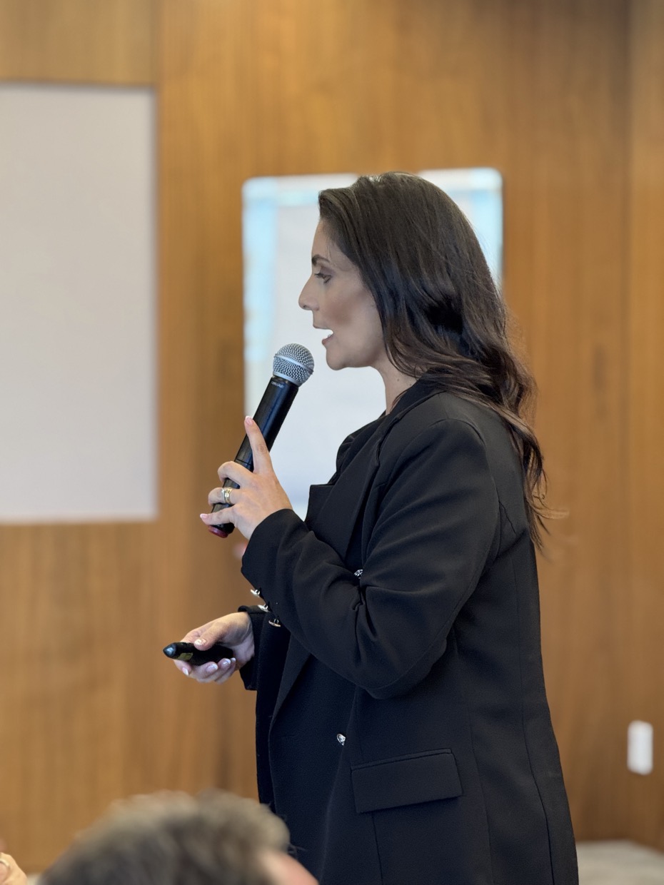 Adriana Ricci falando ao microfone em uma sala de treinamento, de perfil.