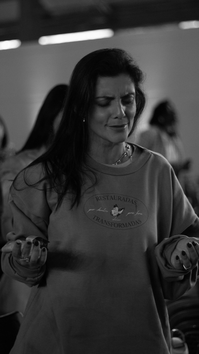Adriana Ricci de olhos fechados, em preto e branco, com as duas mãos erguidas em postura de oração.