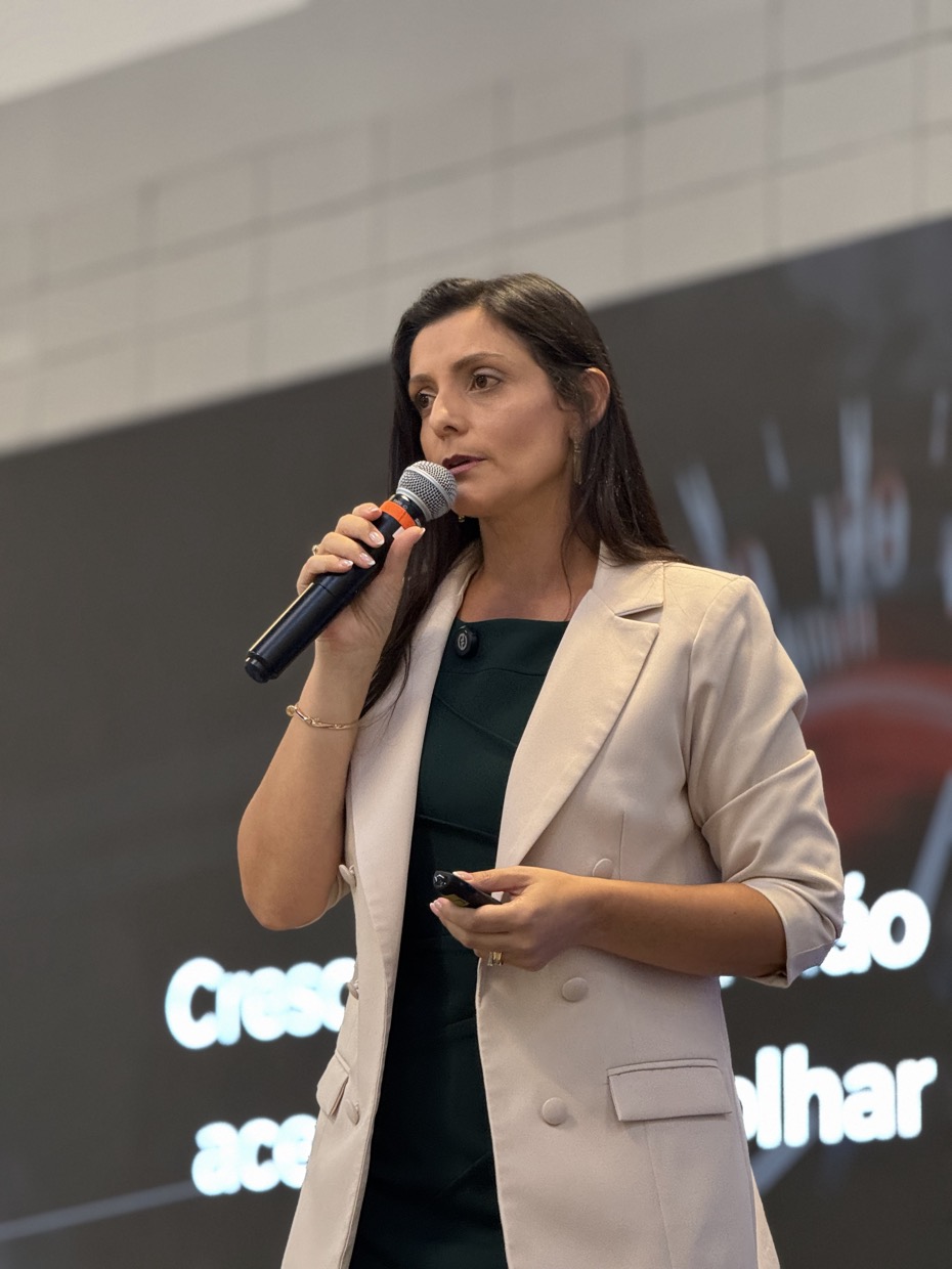 Drica Ricci falando ao microfone diante de um telão escuro, de blazer claro, com o controle da apresentação na mão.